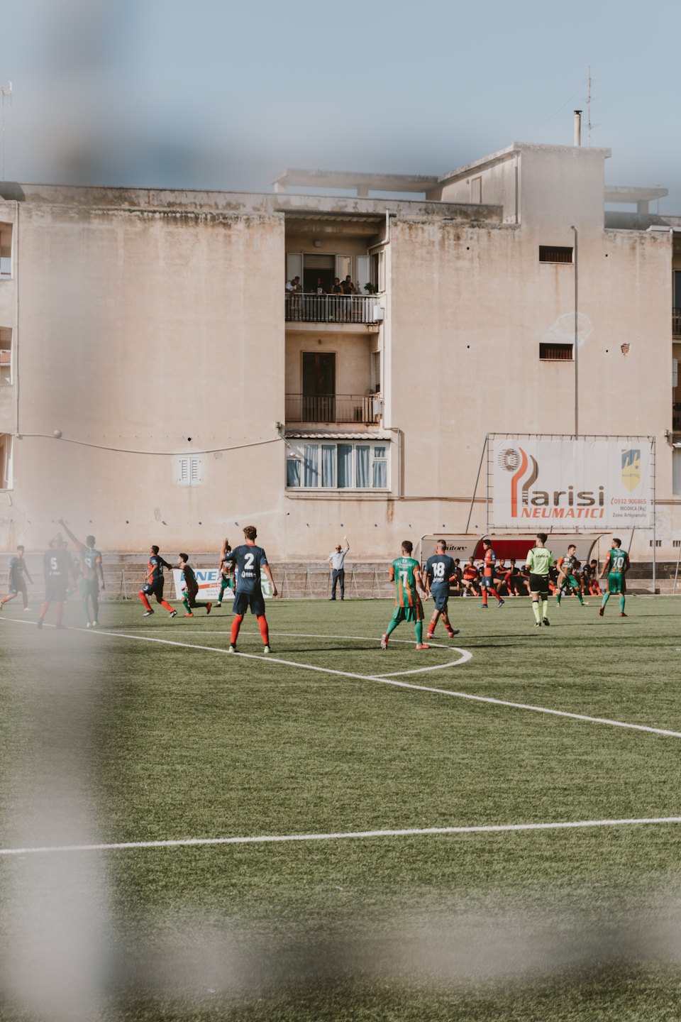 Soccer players compete on an artificial turf field near an apartment building.